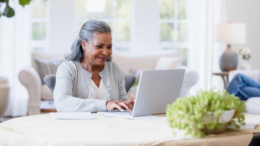 senior woman sitting at her kitchen table using her laptop, researching long term care plans