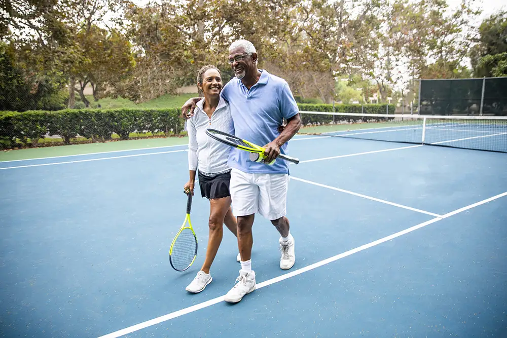 A active senior couple walks arm-in-arm across a tennis court carrying racquets and smiling, illustrating the vibrant lifestyle for The Winsberg residents in luxury senior living in Florida.