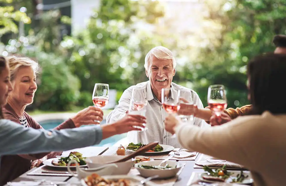 A senior man and his companions toast glasses of rose wine around a filled outdoor dining table, showcasing how seniors at The Winsberg enjoy luxury senior living in Florida.