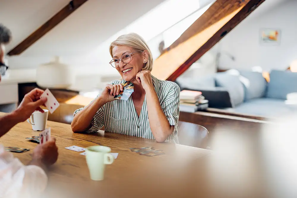 A senior woman wearing glasses smiles warmly while playing a game of cards at a wooden table in a sunlit room, showcasing how The Winsberg at Green Cay residents enjoy relaxing activities in maintenance-free senior living.